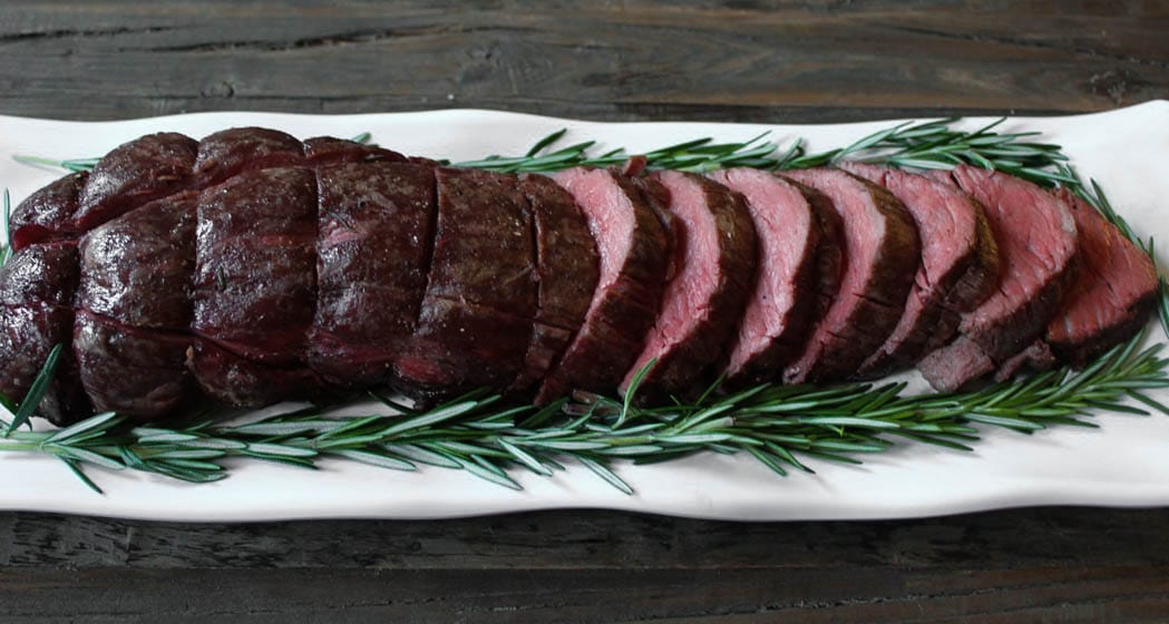 Whole beef tenderloin partially sliced on a platter with fresh rosemary sprigs for presentation.