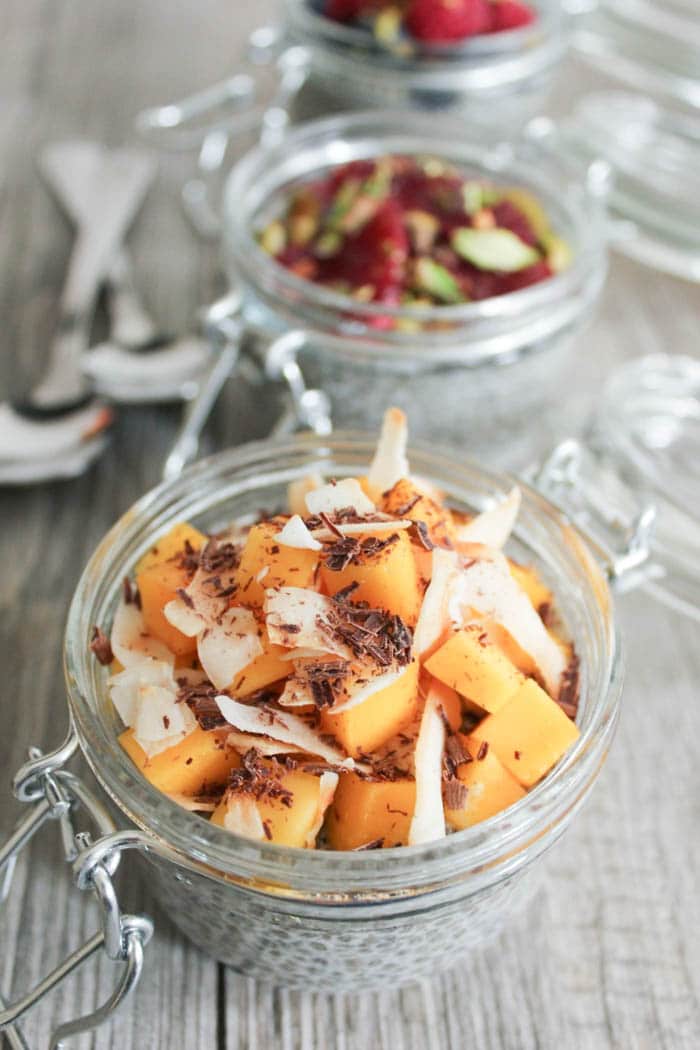 Two jars of chia seed pudding, one topped with mango, coconut, and shaved chocolate, and one topped with blood orange segments and pistachios.