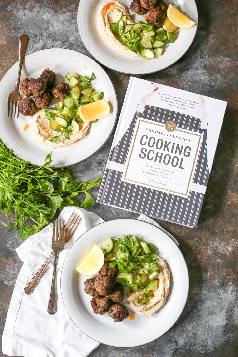 Overhead shot of three plated servings of lamb kofta with a side of hummus and herb salad next to a copy of the Haven's Kitchen Cooking School cookbook.