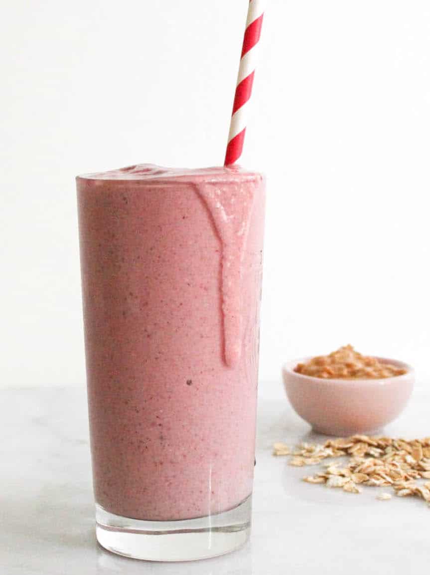 Light red PB&J smoothie in a tall glass with a red and white spiral straw and a bowl of peanut butter in the background.