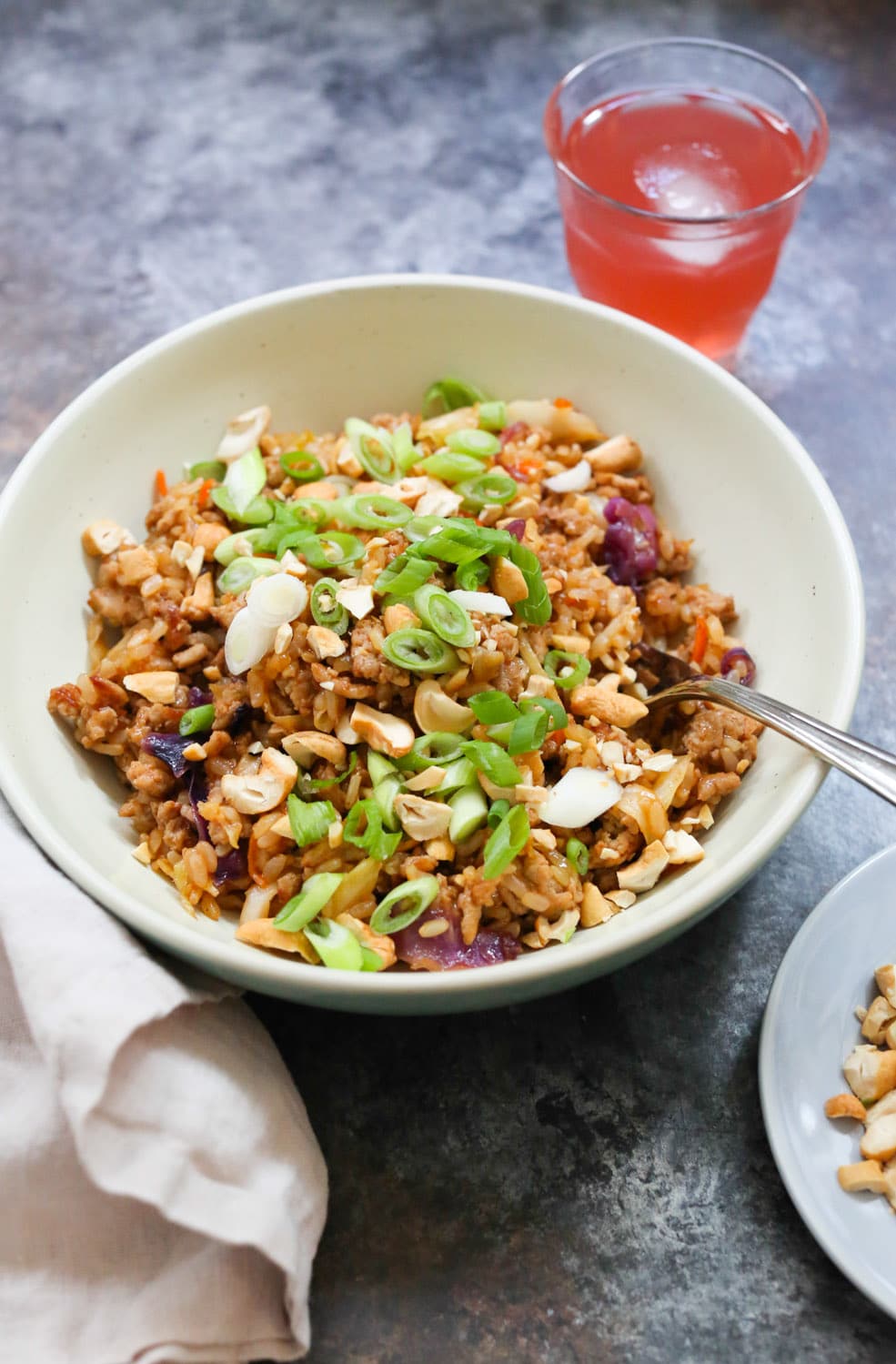 Close-up of one stir-fried turkey and rice bowl topped with scallions and cilantro with a small cup of iced kombucha.